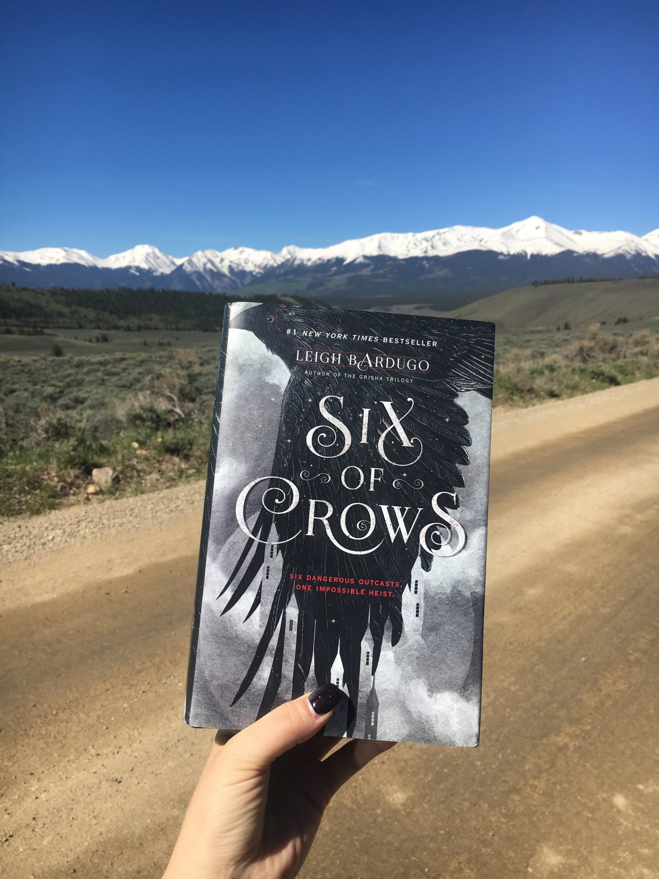 Hardcover copy of Six of Crows in front of a winding dirt road. Background is snowy mountains and bright blue sky