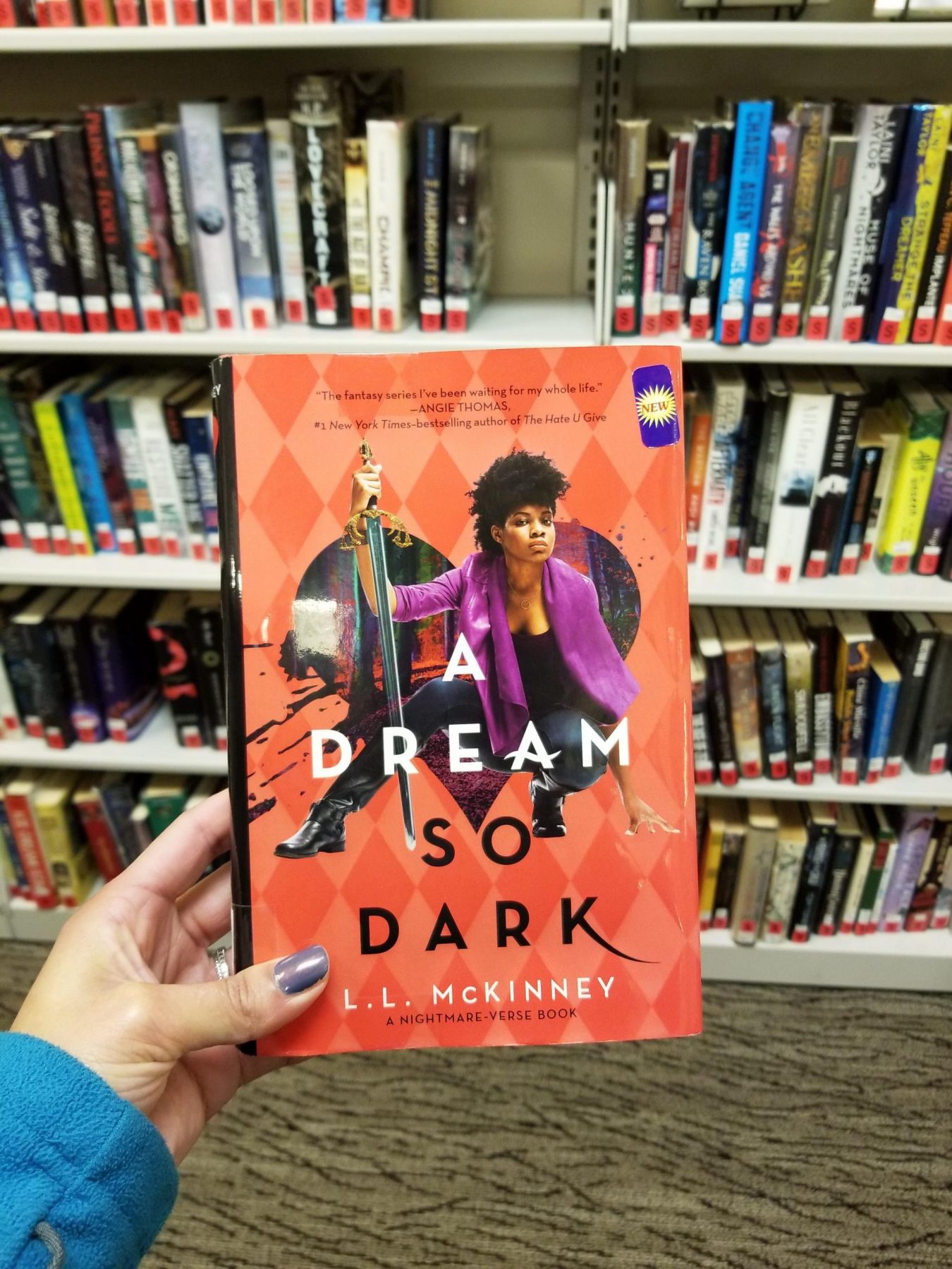 Light-skinned brown hand holds novel A DREAM SO DARK. Shelves of library books in the background.