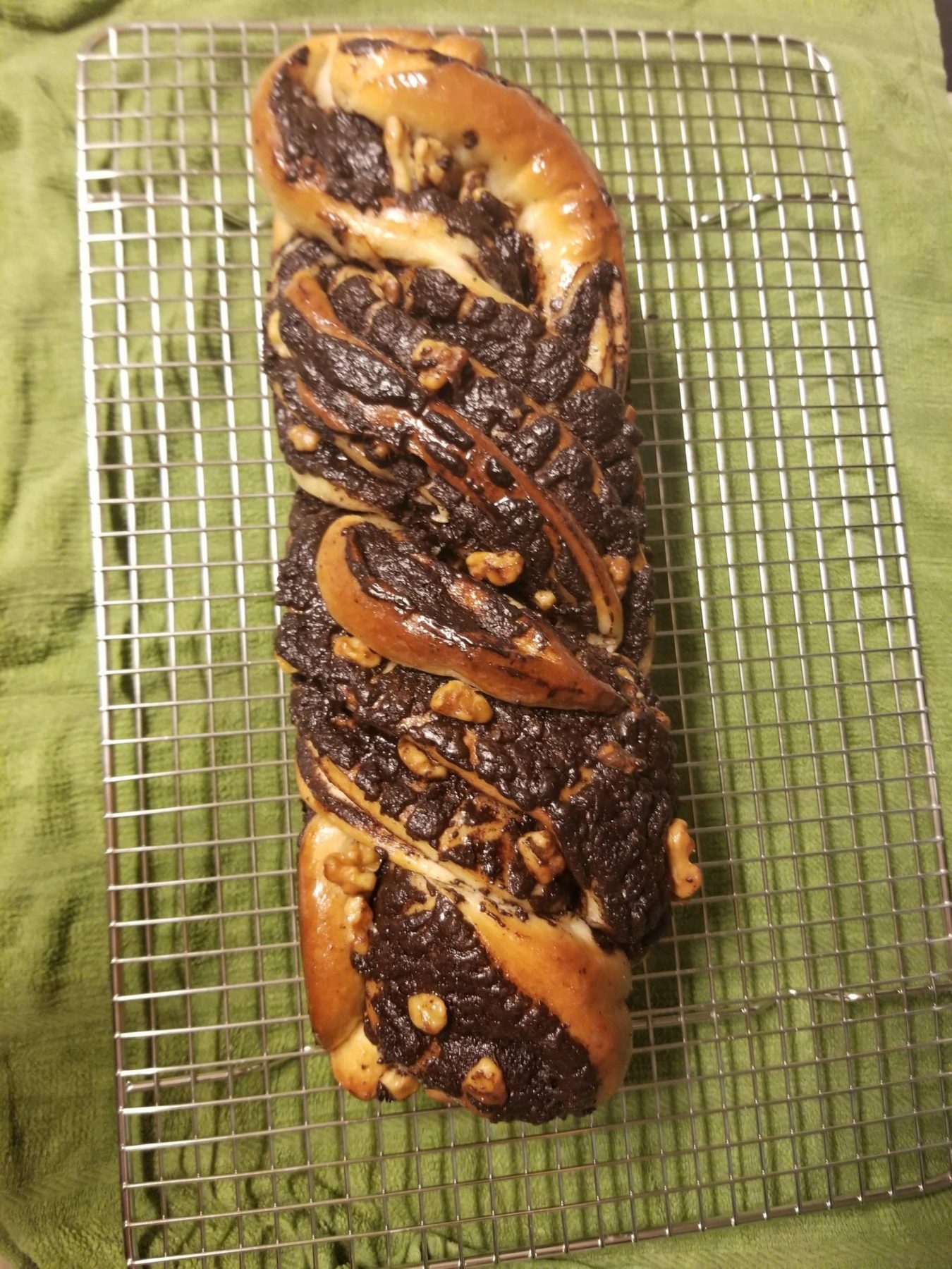 A golden brown chocolate & walnut babka loaf rests on top of a wire cooling rack and sage green tea towel