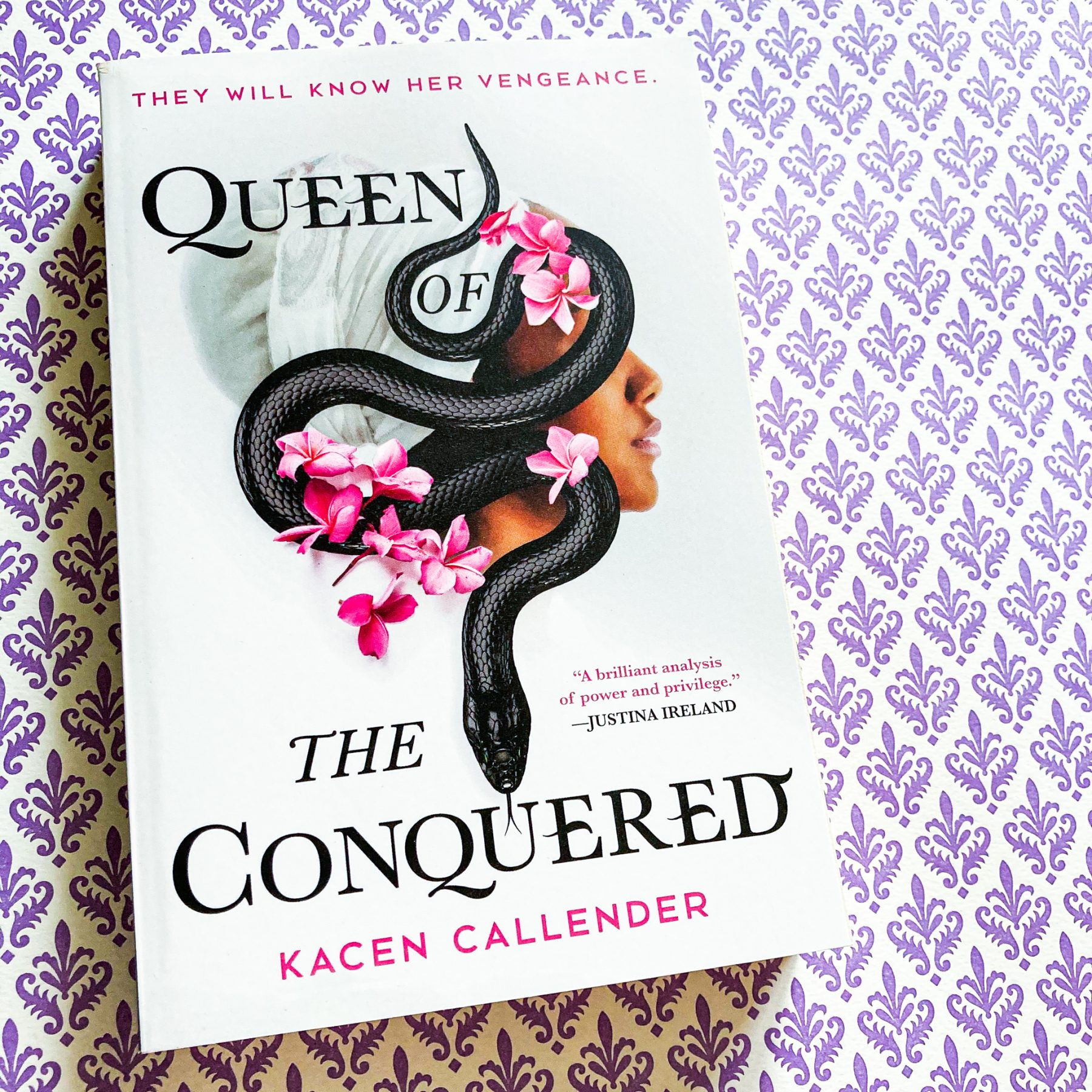 paperback copy of Queen of the Conquered on top of a purple and white decorative paper