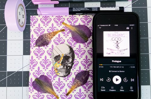 light purple stationary items, pens, Washi tape beside an iPhone with the screen showing The Burning God on audiobook