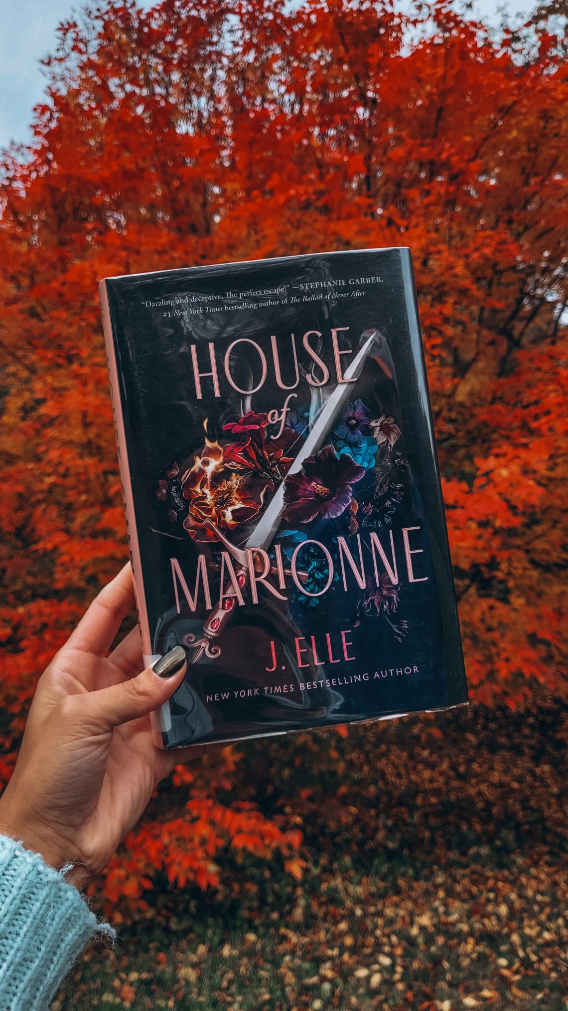 A light brown hand with black nail polish holds up a library copy of House of Marionne in front of a blazing red tree
