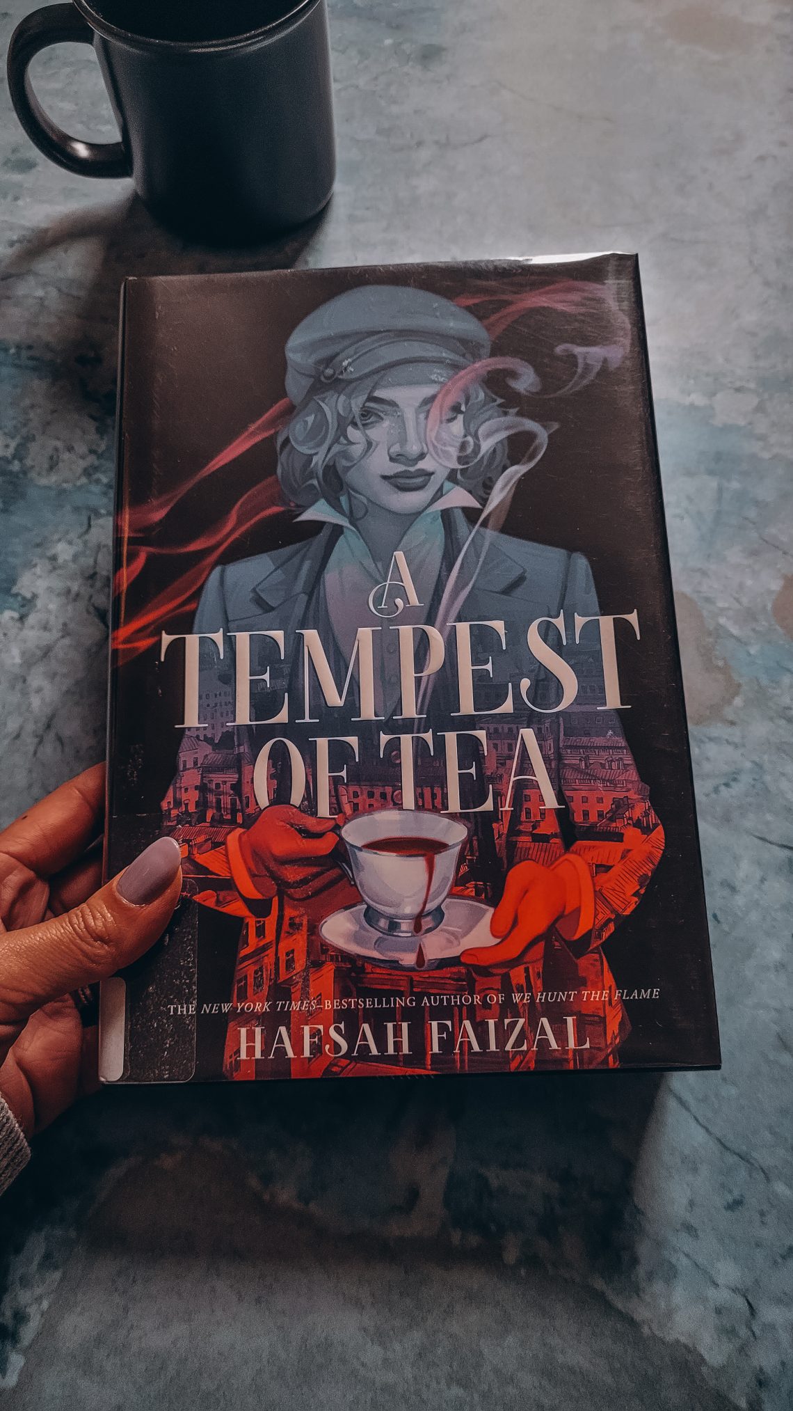 book, A Tempest of Tea, held in a brown hand with a black mug in the background
